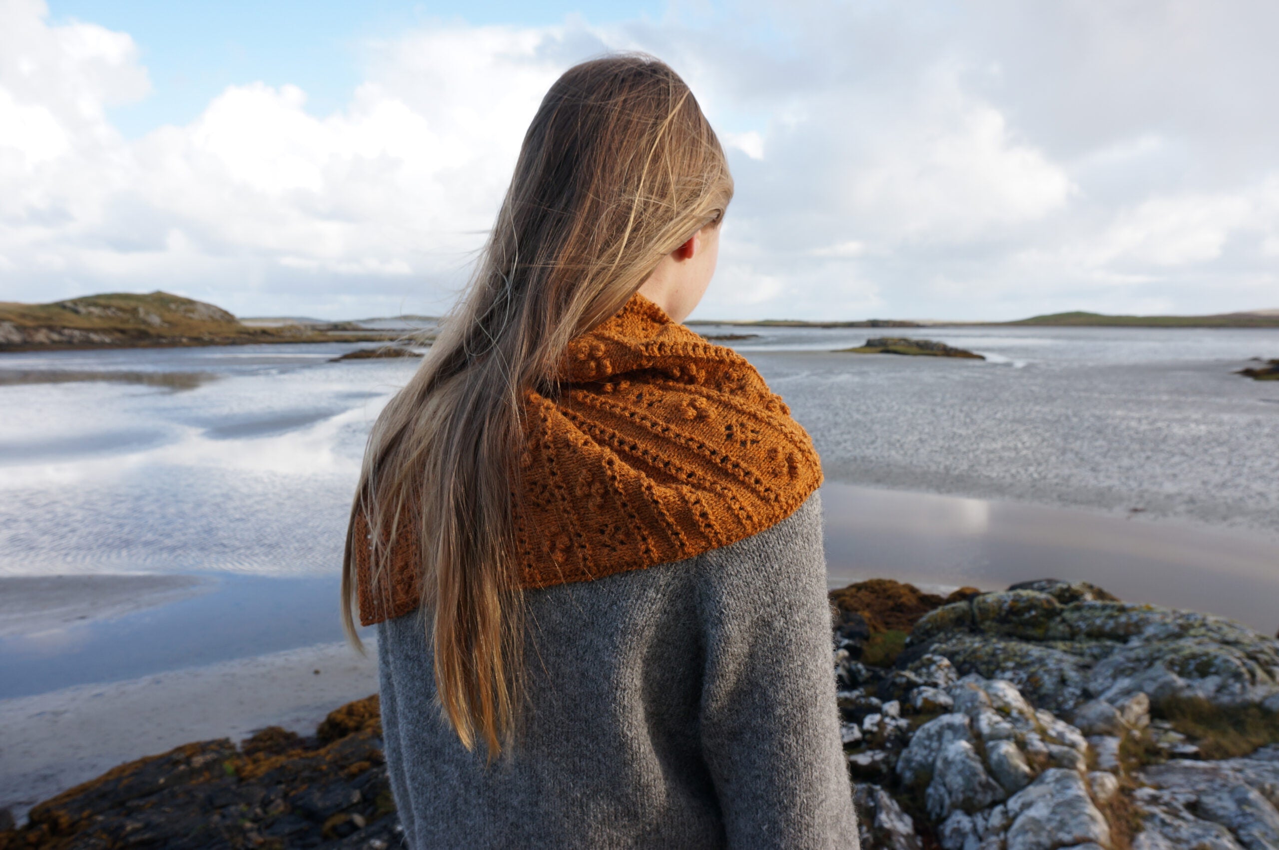 Machair Wildflower Shawl in orange yarn with lace eyelets and raised bobbles worn over a grey jumper by someone on the coast