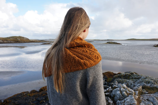 Machair Wildflower Shawl in orange yarn with lace eyelets and raised bobbles worn over a grey jumper by someone on the coast