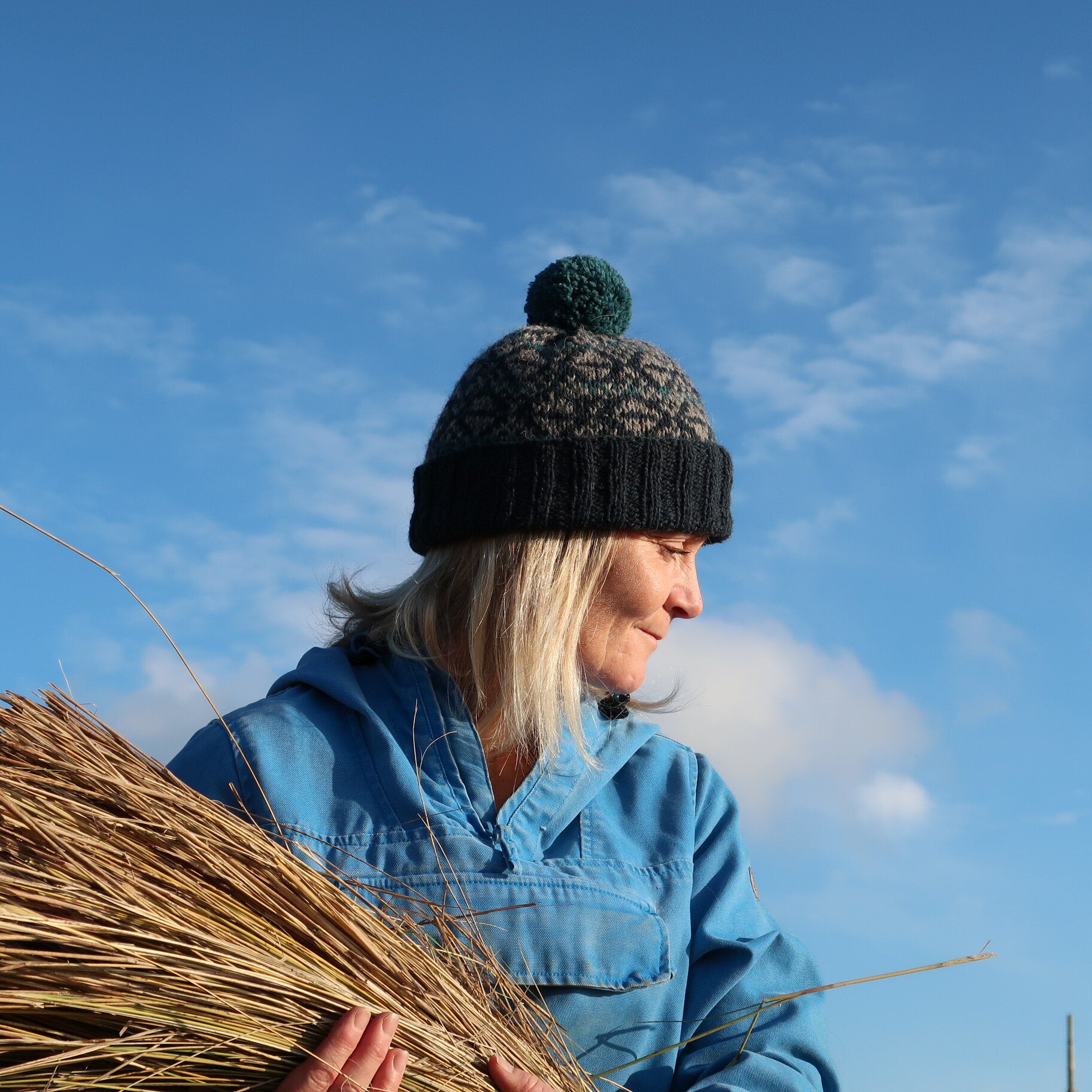 Thatching Hat, beanie with ribbed turn up, colourwork knitting and a bobble worn by Meg outside while holding marram grass