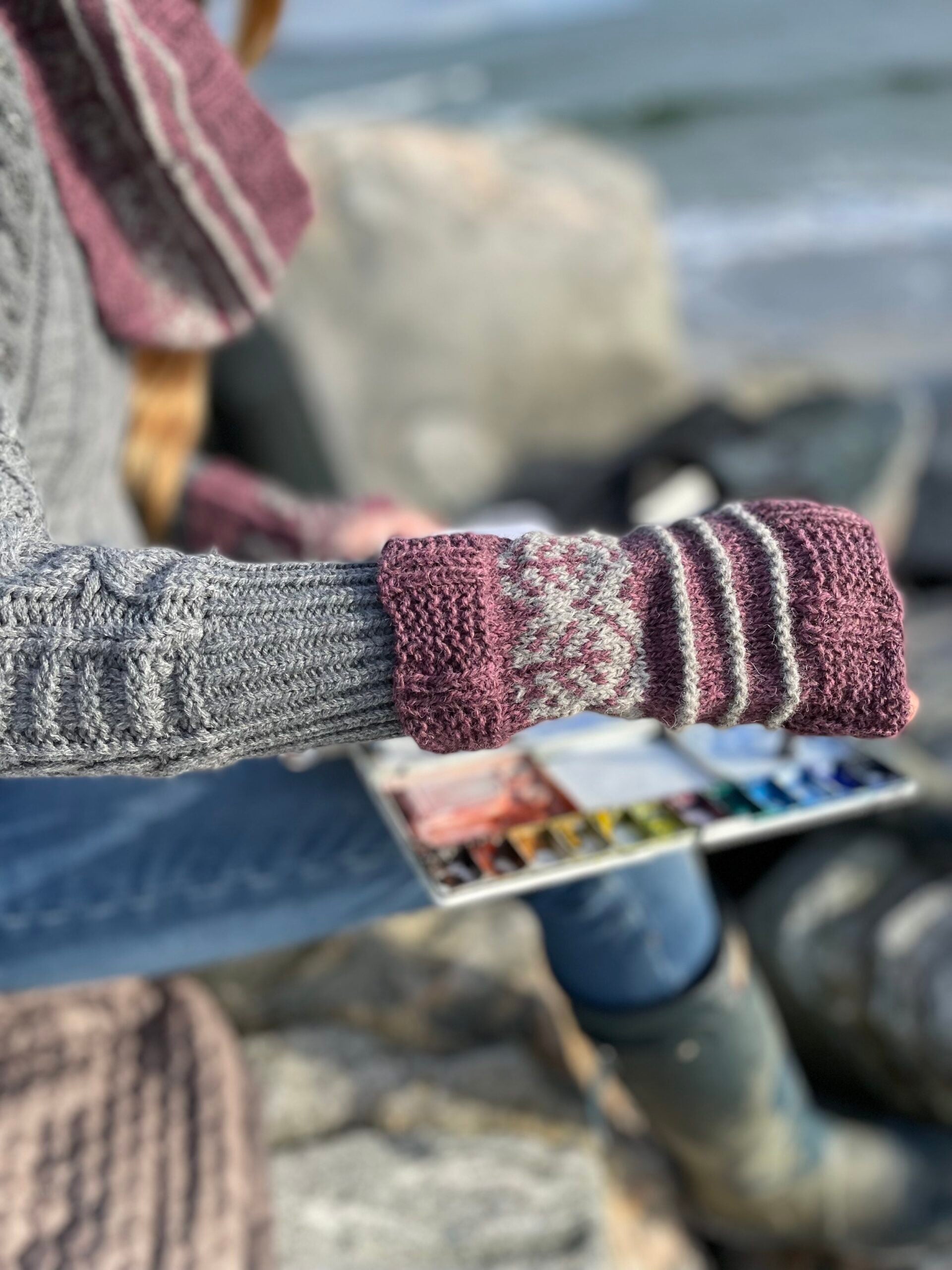Person painting on rocks by a body of water wearing Pottery Tree Wrist Warmers and Cowl Pattern by Emma Vining. They have a knitted motif of a tiny tree within a white oval and white stripes.