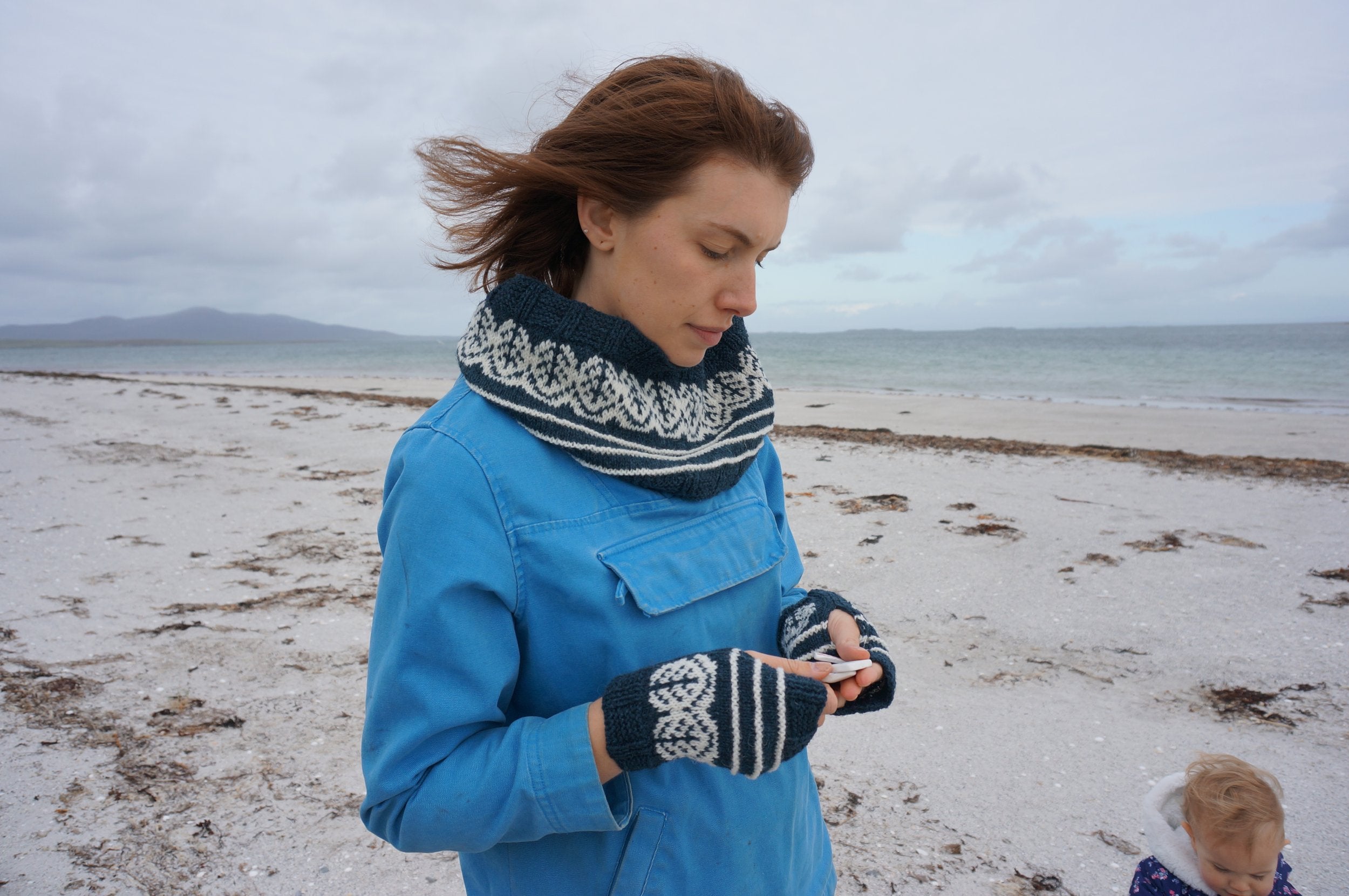Woman in blue coat standing on a beach with the sea in the background. They're wearing Pottery Tree Wrist Warmers and Cowl Pattern by Emma Vining. They have a knitted motif of a tiny tree within a white oval and white stripes. the rest of the cowl and wrist warmers are blue.