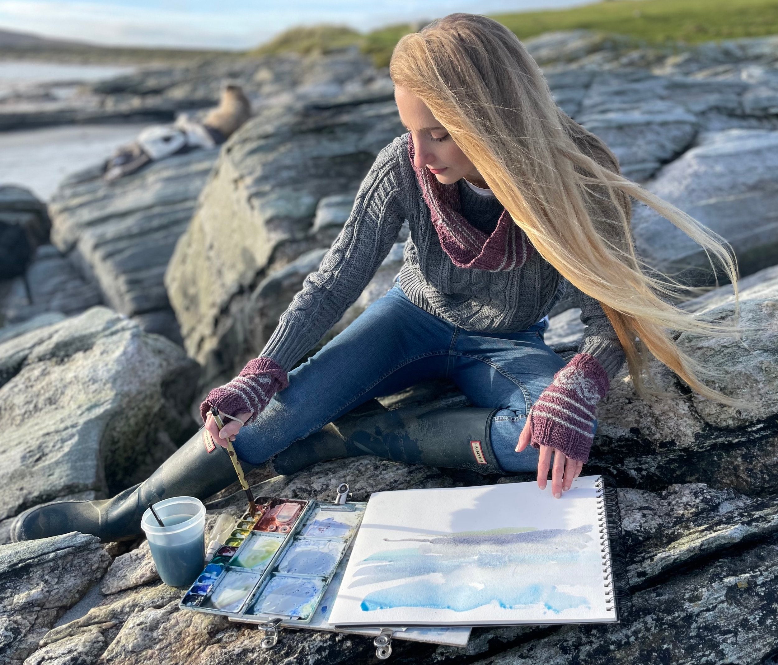 Person painting on rocks by a body of water wearing Pottery Tree Wrist Warmers and Cowl Pattern by Emma Vining. They have a knitted motif of a tiny tree within a white oval and white stripes.
