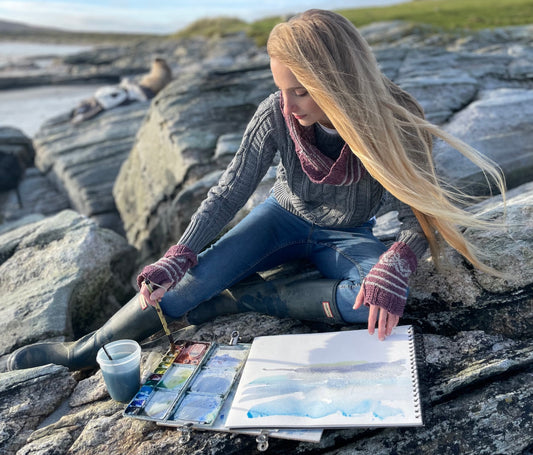 Person painting on rocks by a body of water wearing Pottery Tree Wrist Warmers and Cowl Pattern by Emma Vining. They have a knitted motif of a tiny tree within a white oval and white stripes.