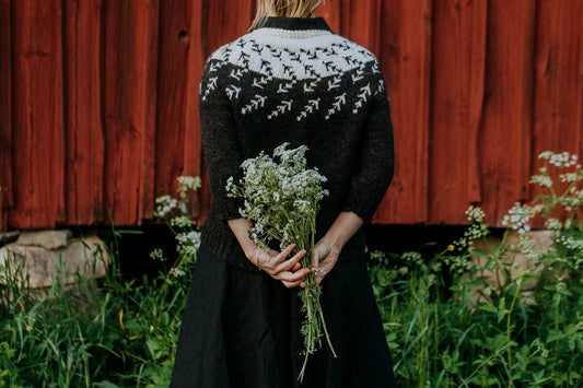 Salix Alba sweater with a circular yoke featuring a pattern resembling arrows or branches. Worn outside while the model holds flowers